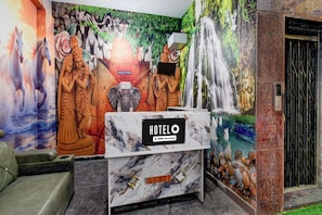 Un mostrador de recepción de hotel con encimera de mármol, un letrero que indica 'HOTEL' y 'o-v-serviced', y un mural que representa varias figuras y una cascada.