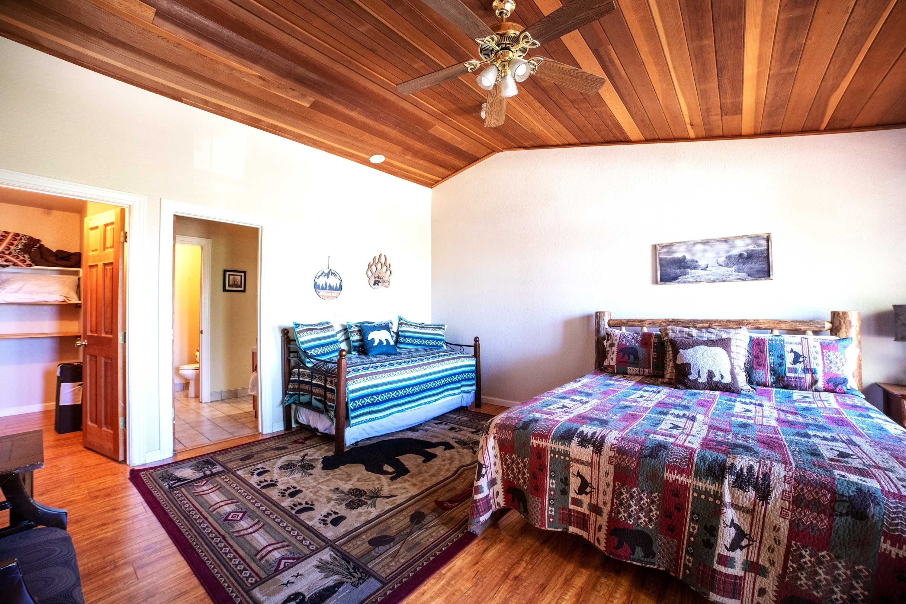Un dormitorio con techo de madera, una cama con ropa de cama estampada, una alfombra con motivos de animales y una puerta que da a otra habitación.
