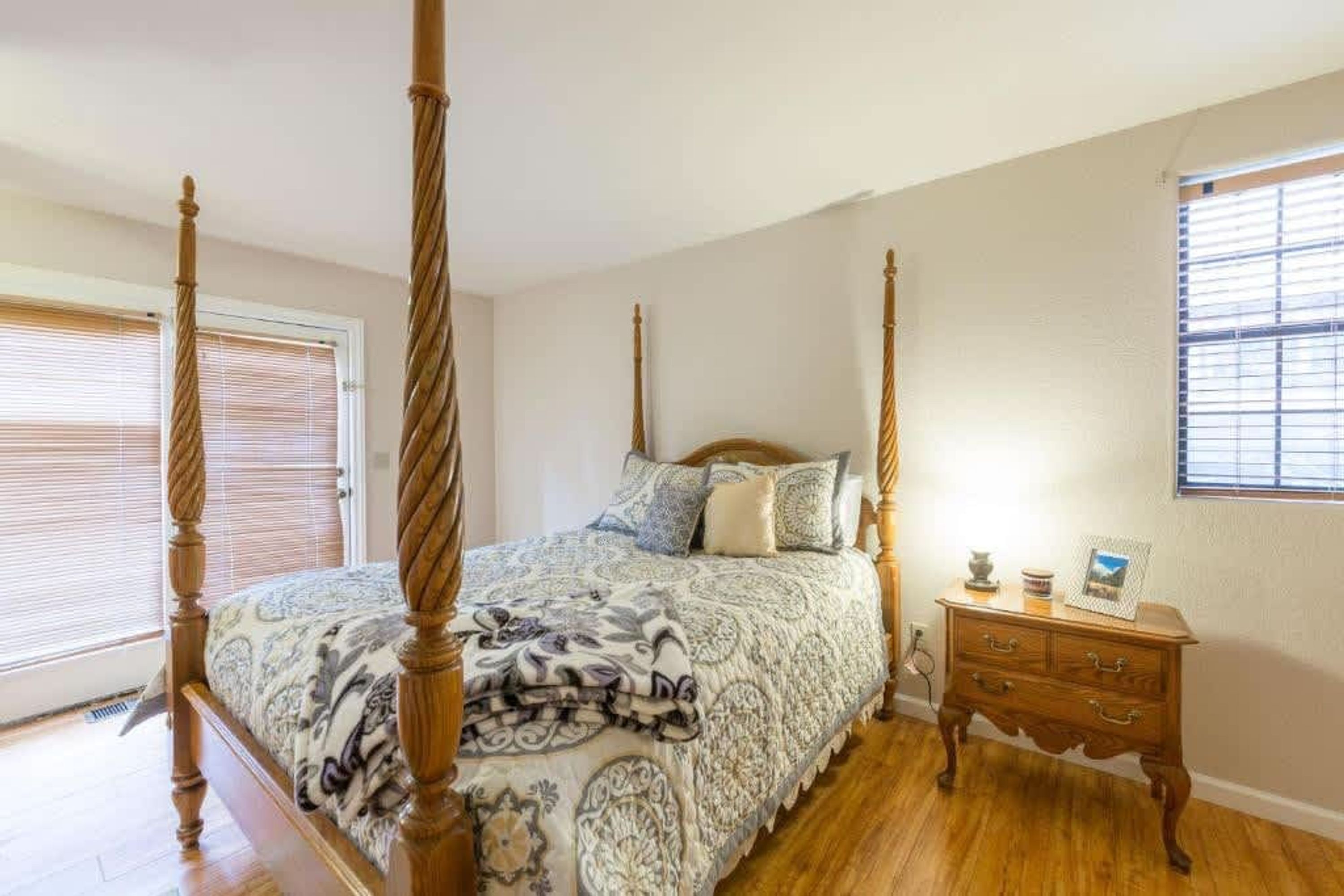 A four-poster bed with a patterned bedspread, a wooden nightstand with drawers, and a window with blinds.