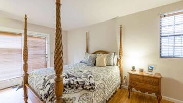A four-poster bed with a patterned bedspread, a wooden nightstand with drawers, and a window with blinds.
