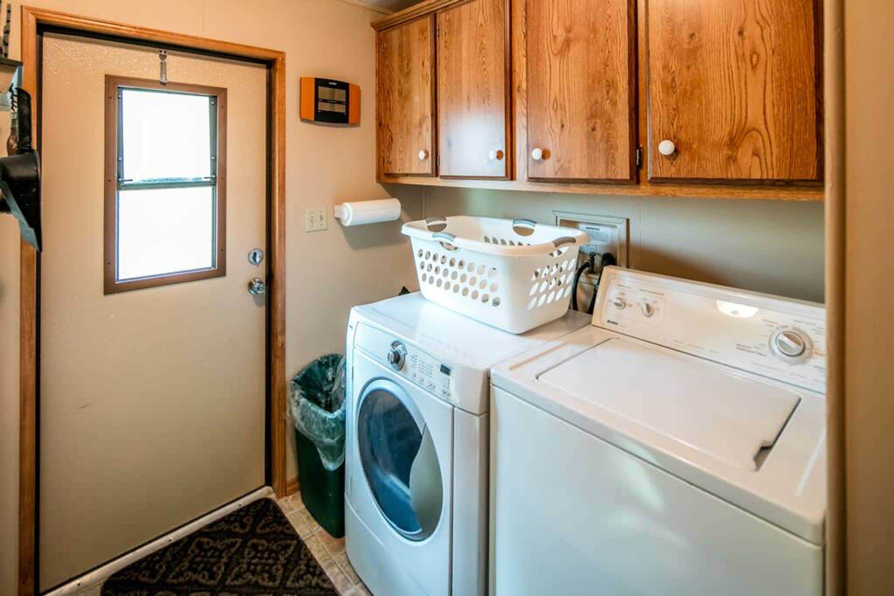 A laundry room with a front-loading washer and dryer, wooden cabinets above, a window, and a trash can.