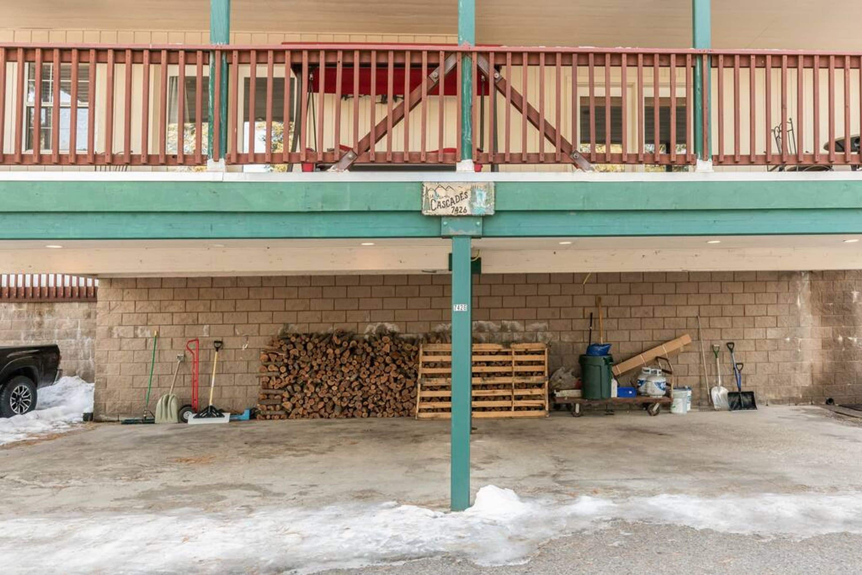 Um edifício com área de estacionamento coberta, lenha empilhada e diversas ferramentas.