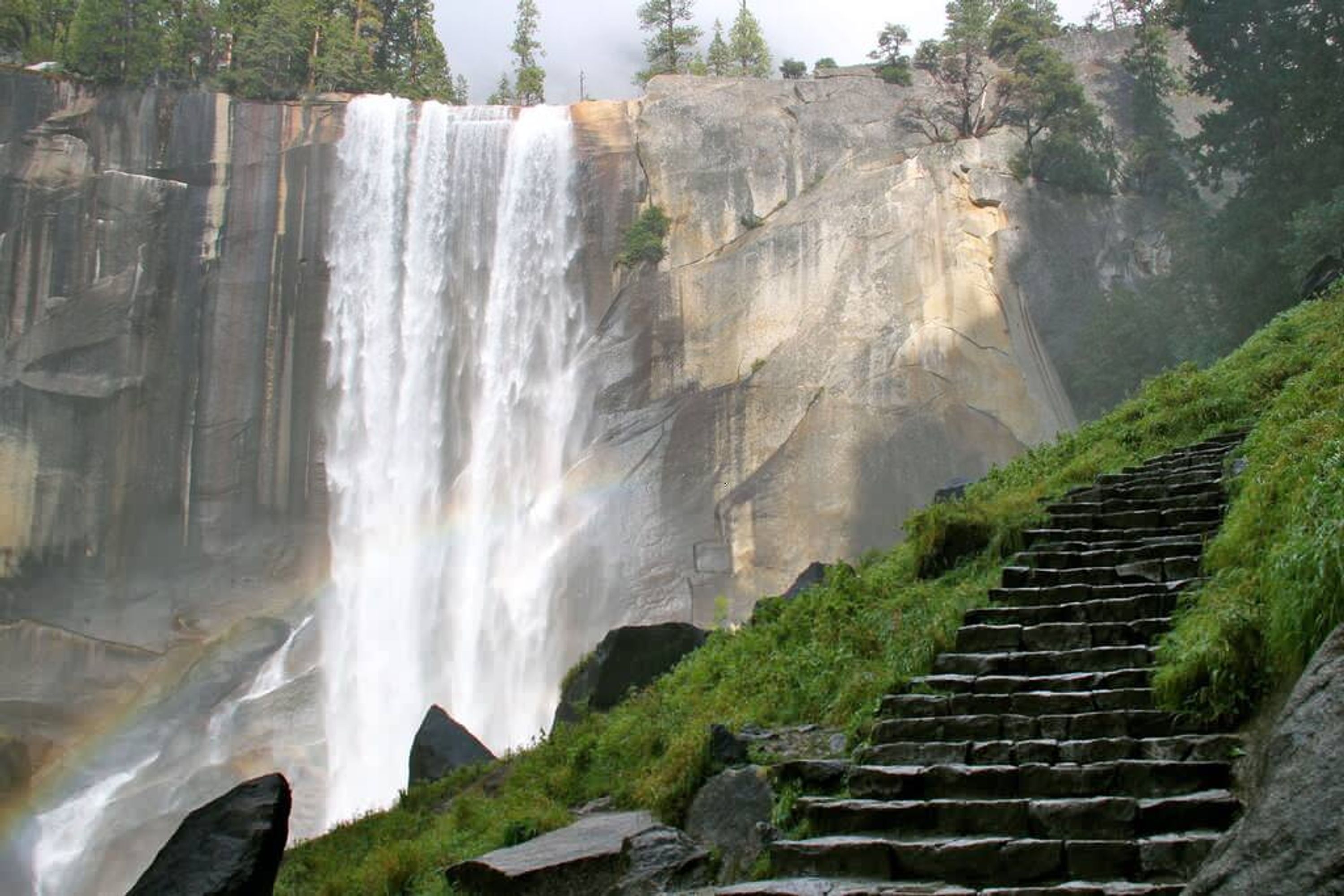 Una cascata che si getta da una scogliera rocciosa, con una scala di pietra che sale verso di essa.