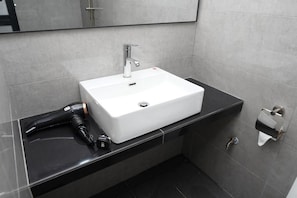 A bathroom with a white sink, a mirror, a black countertop, a hairdryer, and a paper towel dispenser.