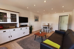 Ein modernes Wohnzimmer mit einer Couch, einem Couchtisch und einem Fernseher. Es gibt einen Essbereich mit Tisch und Stühlen.