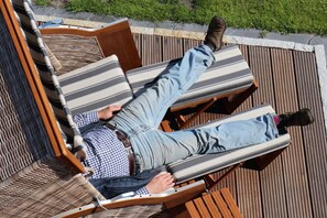 Eine Person entspannt auf einer Holzterrasse, ausgestattet mit gestreiften Kissen, einem karierten Hemd und hellen Hosen.