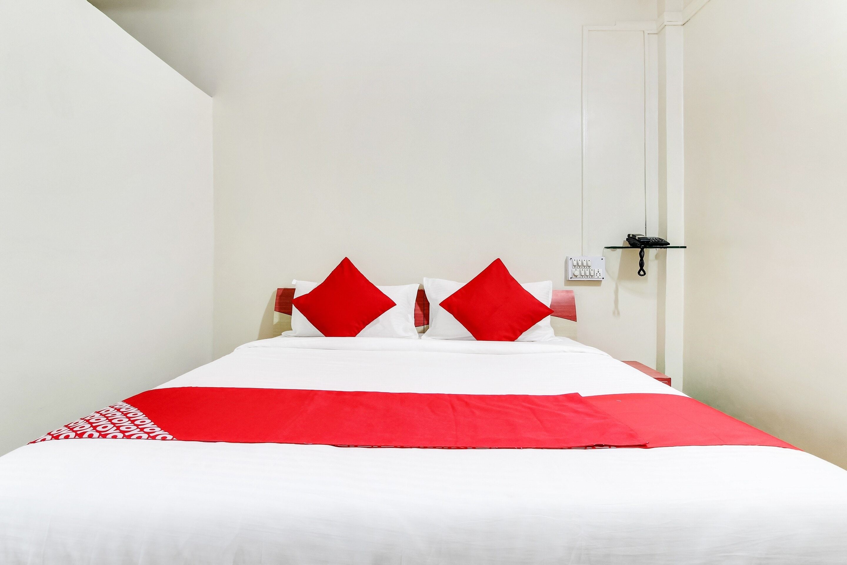 Een hotelkamer met een bed, twee rode kussens, een witte spreideken en een telefoon op een glazen plank.
