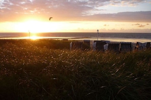 Ein Strand mit Sonnenuntergang, einem grasbewachsen Vordergrund und Liegestühlen.