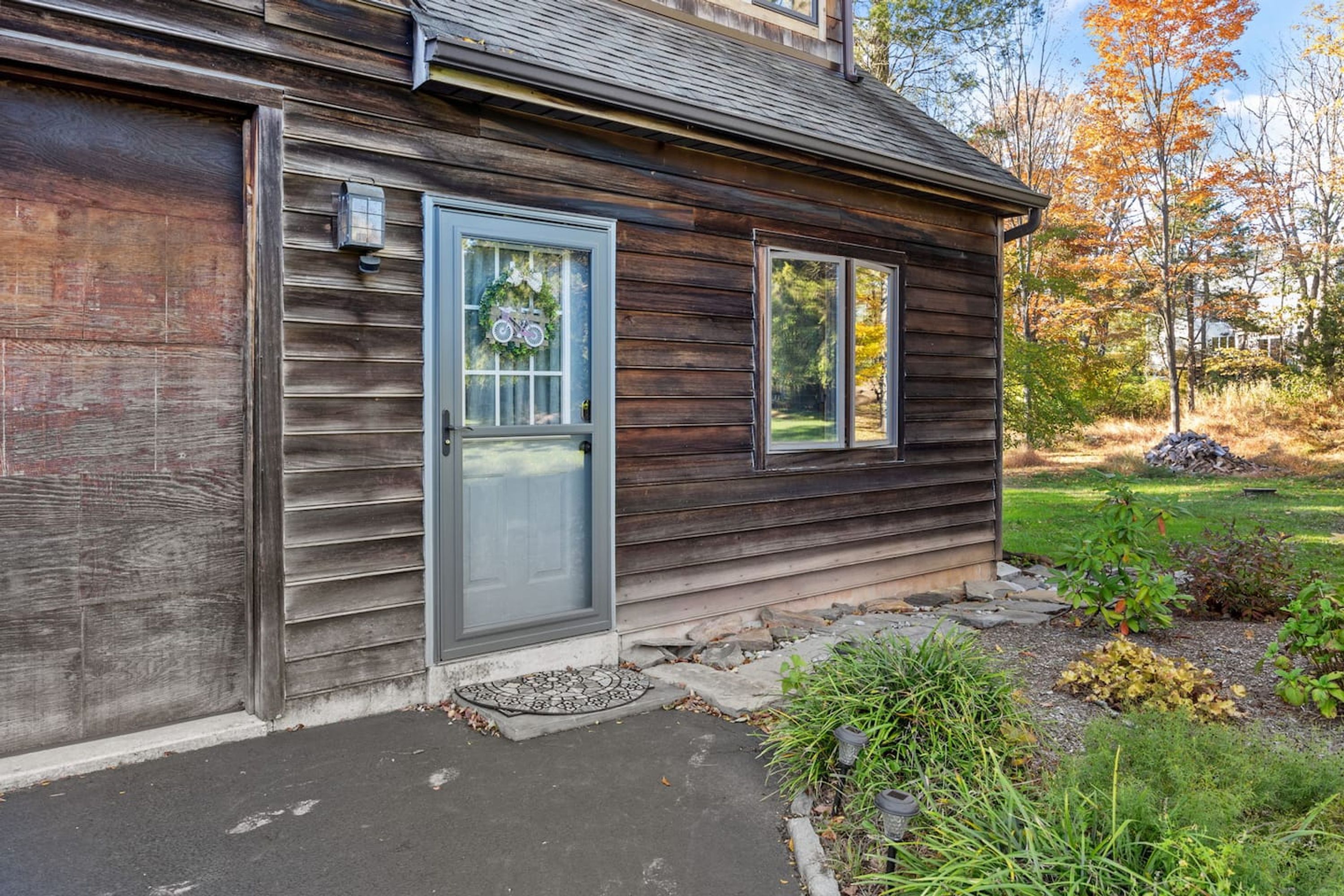A wooden house with a grey door, a window with a wreath, and a small garden with plants and a stone pathway.