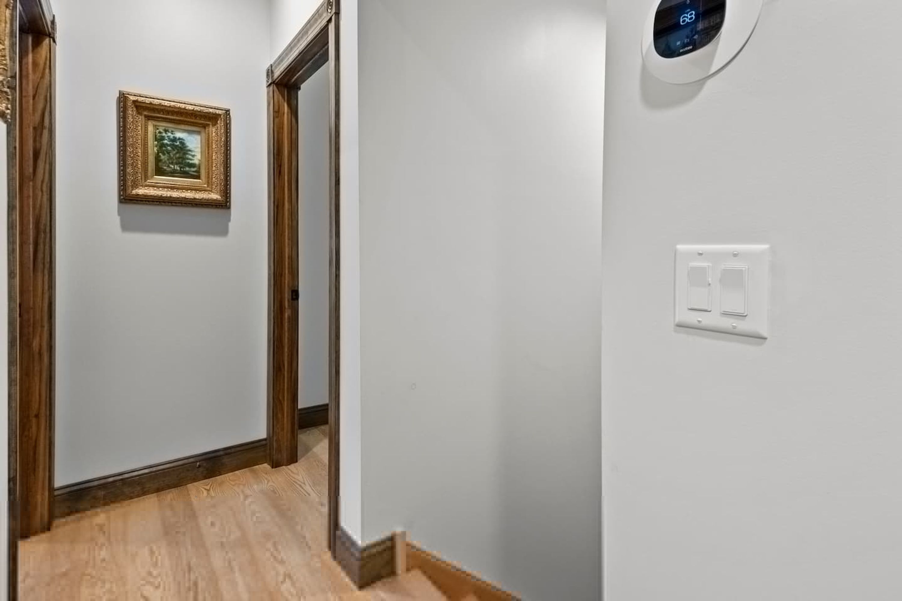 A hallway with a wooden floor, a framed picture on the wall, and a thermostat on the wall.