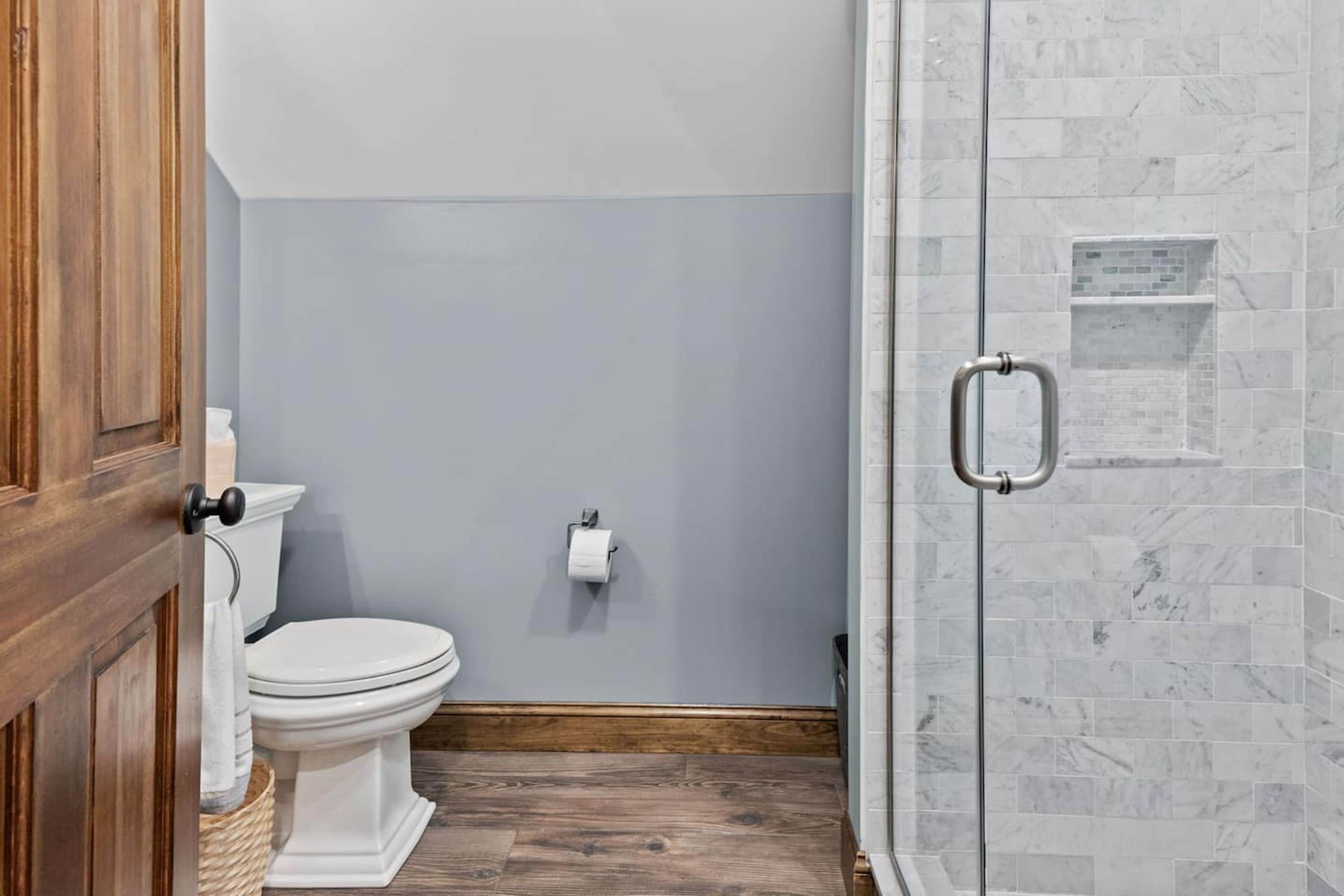 A bathroom with a toilet, a roll of toilet paper, a wooden floor, and a shower with a glass door.