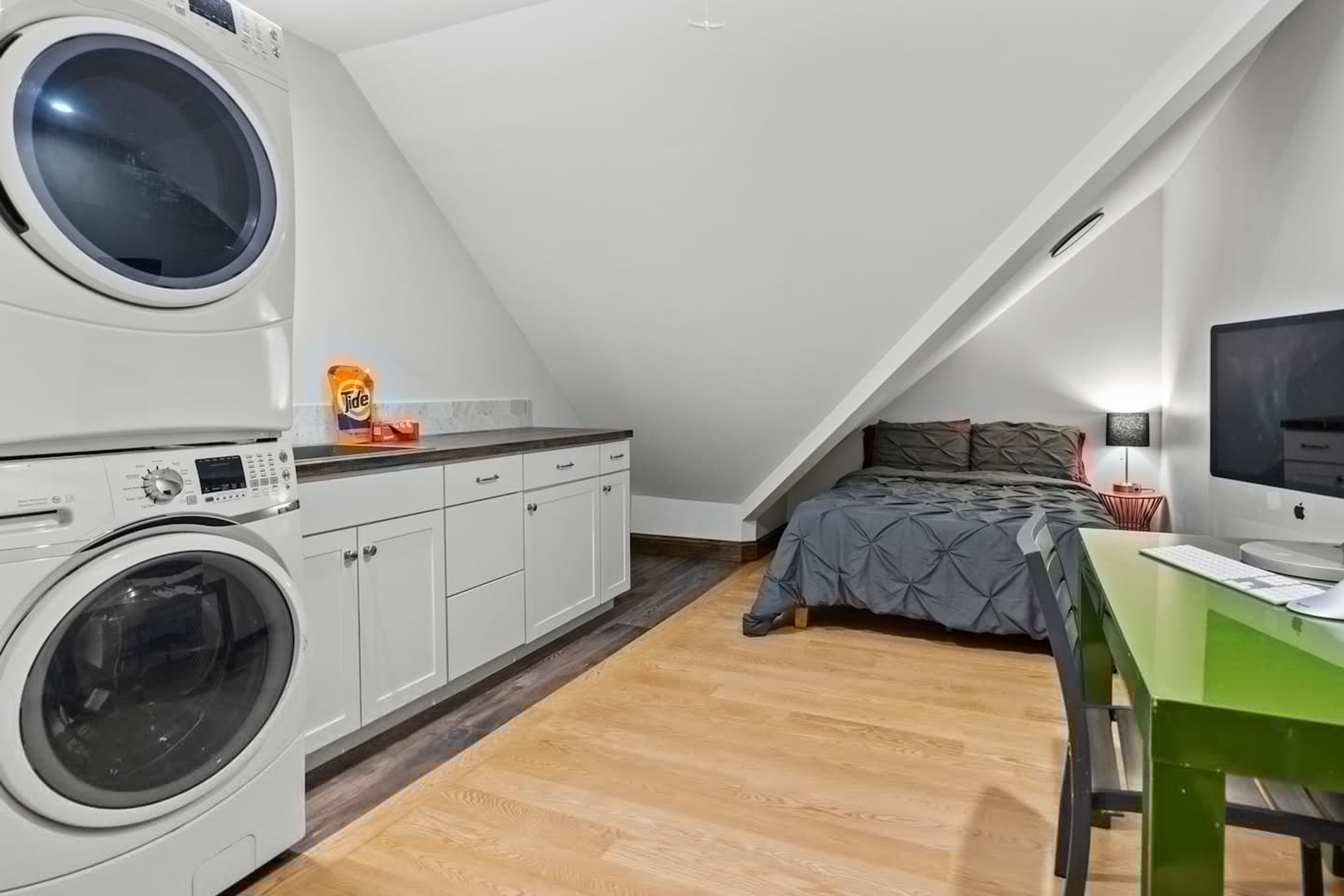 Een kleine kamer met een wasmachine, een bed met een grijze dekbedovertrek, een bureau met een computer en een televisie aan de muur.