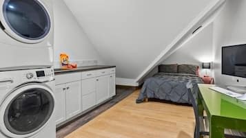 Een kleine kamer met een wasmachine, een bed met een grijze dekbedovertrek, een bureau met een computer en een televisie aan de muur.