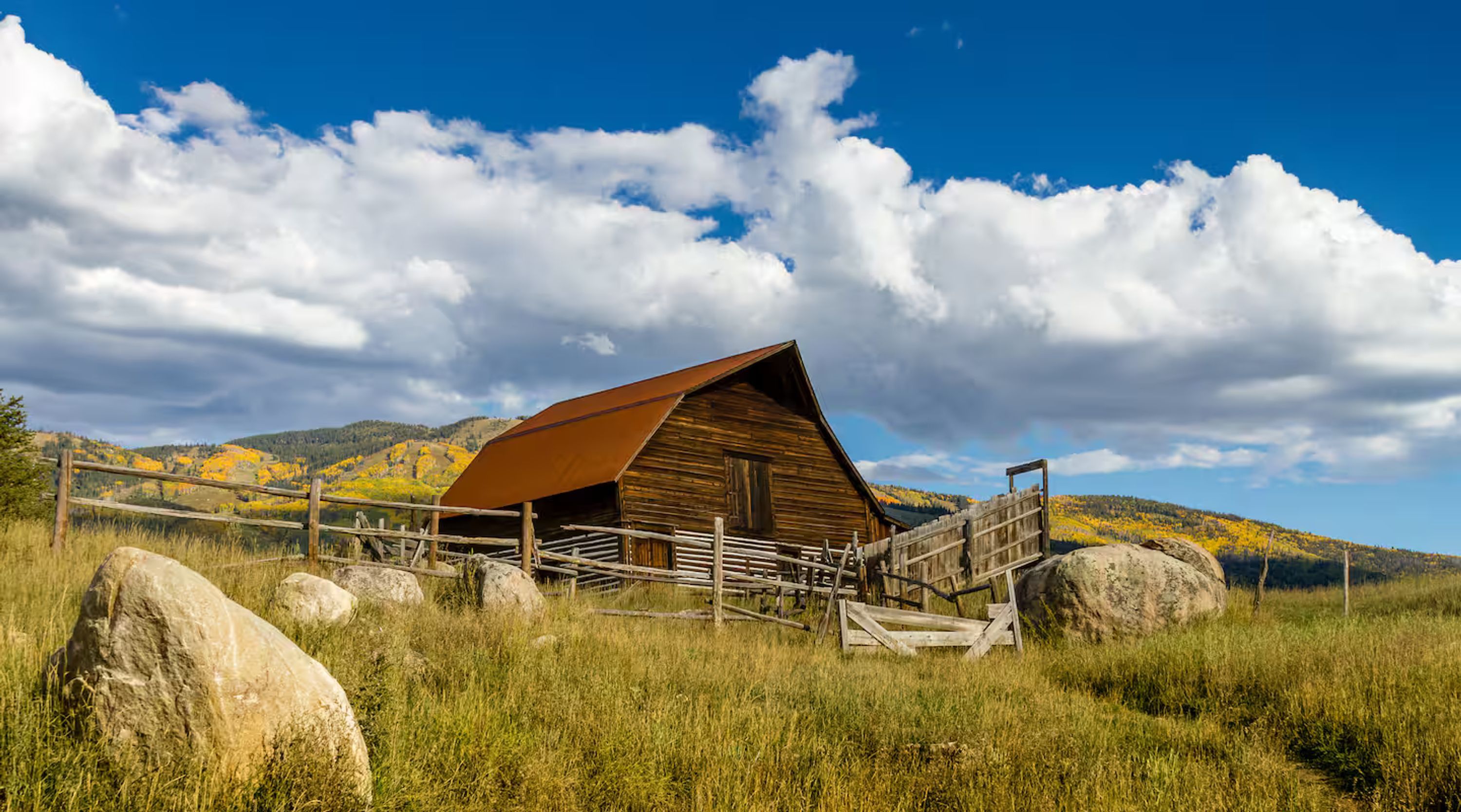 A rustic wooden barn with a metal roof, surrounded by a wooden fence and large rocks, set against a backdrop of hills and a partly cloudy sky.
