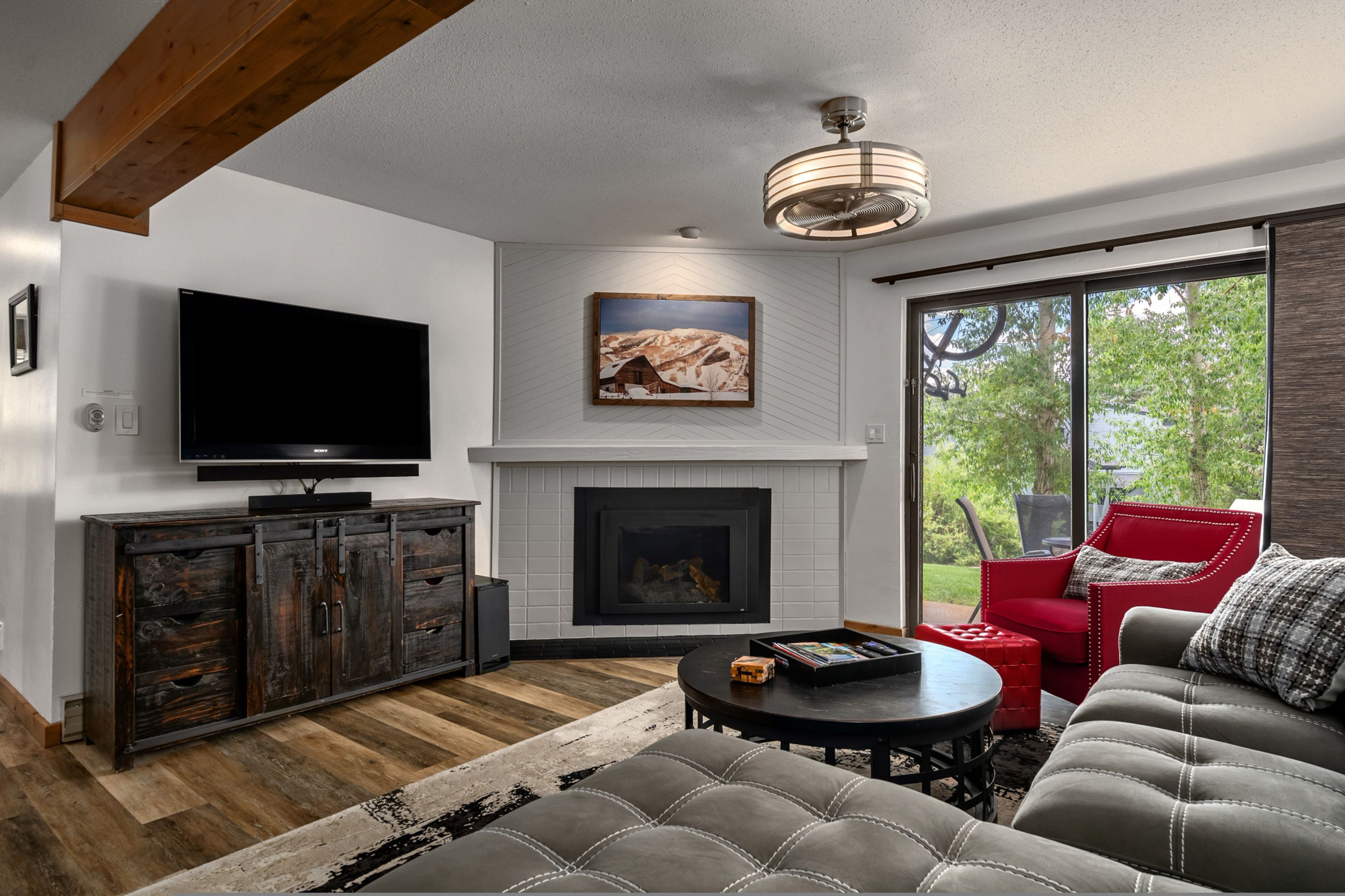 A modern living room with a flat-screen TV, a fireplace, and a large window offering a view of greenery.
