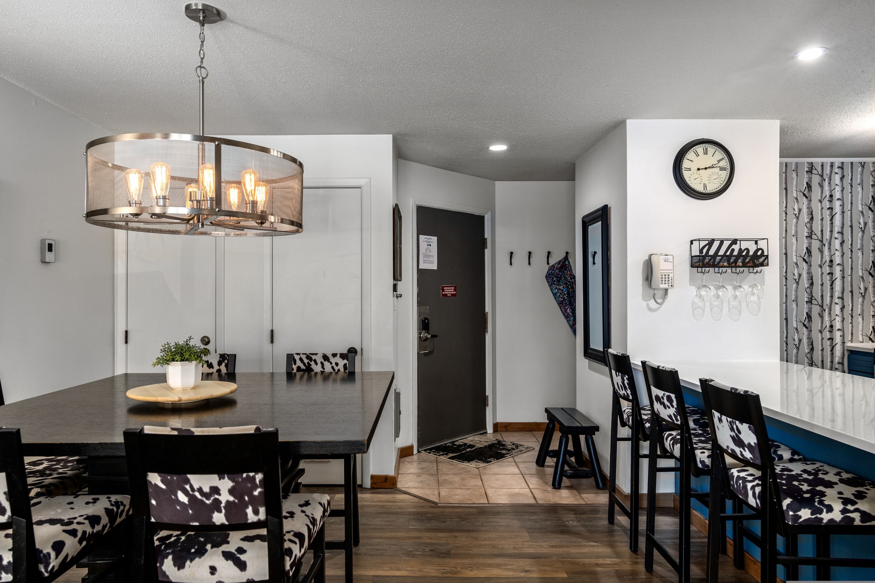 Un comedor con una mesa de madera, sillas con estampado blanco y negro, una lámpara colgante y un reloj de pared.