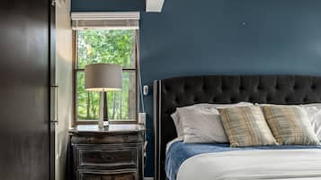 Un dormitorio con pared azul oscuro, un ventanal con vistas a árboles, una mesita de noche con lámpara y una cama con muchos cojines.