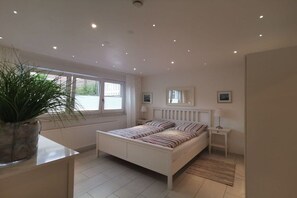 A bedroom with a bed, bedside tables, a radiator, a window, and a plant. - Holiday apartment with 1 bedroom (Norderney)