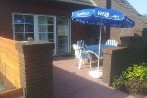 A patio area with a table and chairs, a blue umbrella, and a brick wall. - Holiday apartment with 2 bedrooms (Norden)