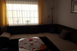 A living room with a sofa, a table with a lace tablecloth, and a window with curtains. - Holiday apartment with 2 bedrooms (Norden)