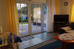 Une pièce comprenant une télévision, un canapé, une table recouverte d’une nappe et une chaise. Une porte-fenêtre coulissante donne accès à un espace extérieur.