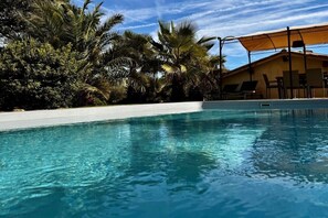 Ein klares Schwimmbecken mit Liegestühlen und Sonnenschirmen, umgeben von Palmen und einem Gebäude.