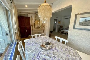 Ein Essbereich mit einem runden Tisch, weißen Stühlen, einer Chandelierleuchtre und einer gemusterten Tischdecke.