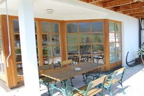 Eine überdachte Terrasse mit Holztisch und Bänken, großen Glasüren und Blick ins Freie.