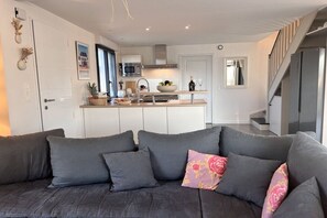 A modern living room with a grey sofa, a kitchen in the background, and a staircase leading to an upper level. - Villa in Brignogan Plages with barbecue (Brignogan Plages)