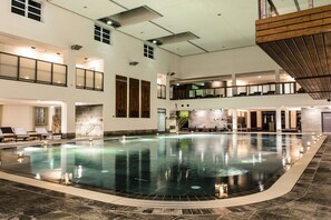 Una amplia piscina interior con suelo de baldosas, rodeada de sillones de descanso y una estructura de techo de madera.