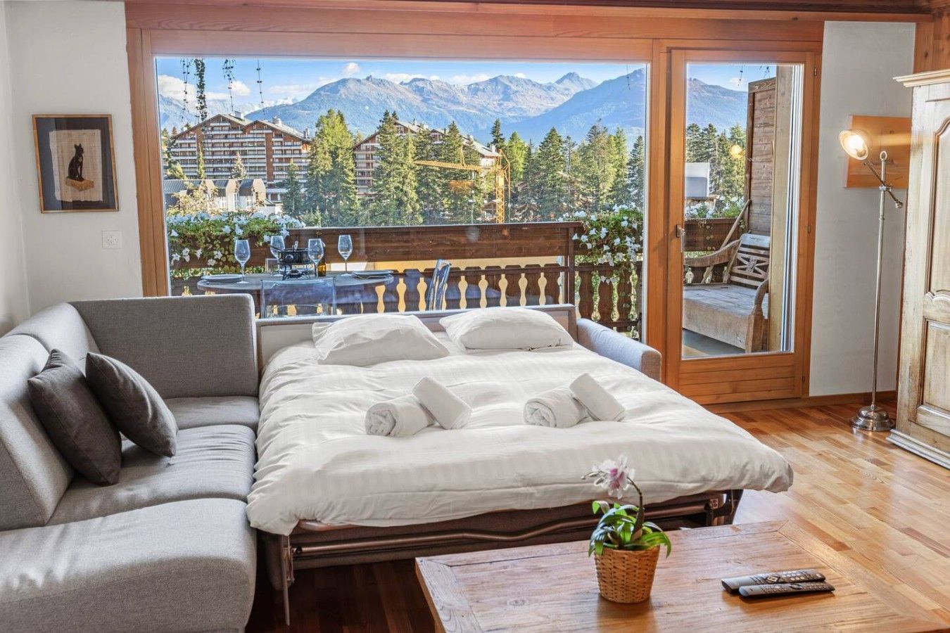 Une chambre d’hôtel avec un lit, un canapé, un plancher en bois, un balcon avec vue sur les montagnes et une télécommande posée sur une table.