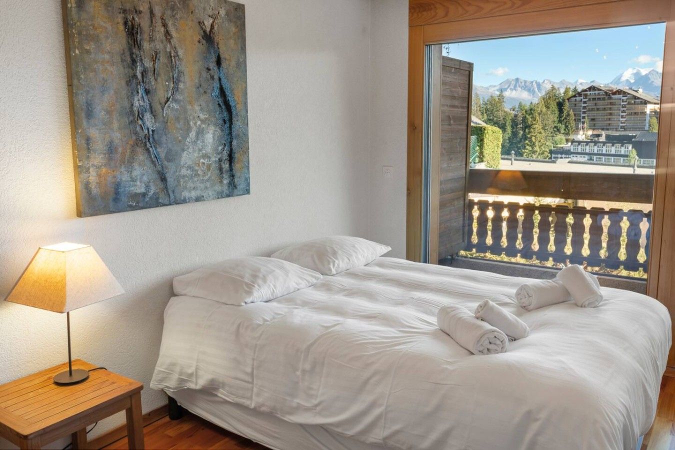 Une chambre à coucher avec un lit, une table de chevet, une lampe et un tableau. Il y a une fenêtre donnant sur des immeubles et des arbres.
