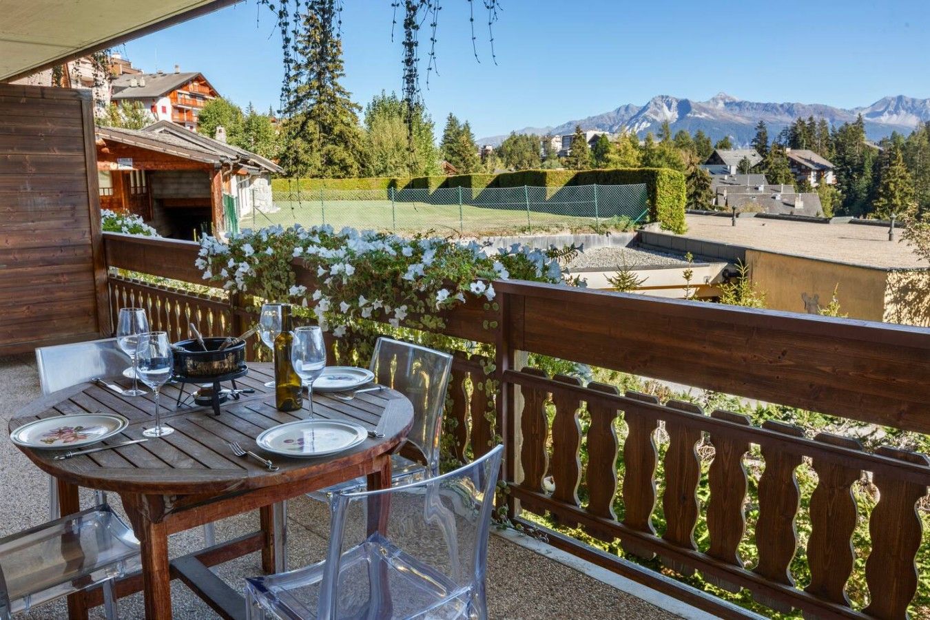An outdoor dining area with a wooden table and chairs, a wine bottle, and a view of mountains in the background.