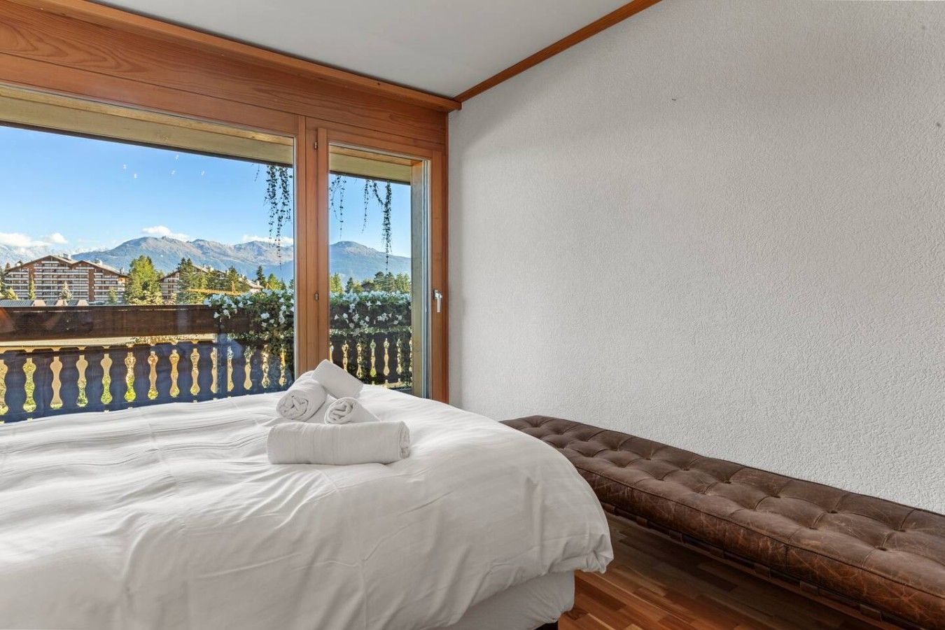 A bedroom with a bed, a bench, and a balcony with a view of mountains and buildings.