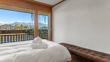 A bedroom with a bed, a bench, and a balcony with a view of mountains and buildings.