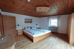 A bedroom with a wooden ceiling, a bed with patterned bedding, a nightstand, a window with curtains, and a radiator. - Holiday apartment with 1 bedroom (Schönau am Königssee)