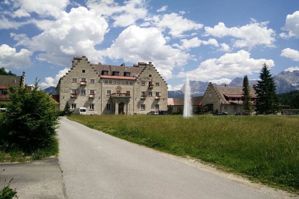Un grand bâtiment en pierre, doté de nombreuses fenêtres et d’une entrée centrale, entouré d’un champ verdoyant et bordé d’une route pavée.