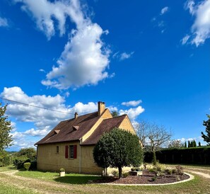 Une maison de plain-pied avec un toit en pente, un petit jardin devant et un ciel bleu dégagé parsemé de nuages.