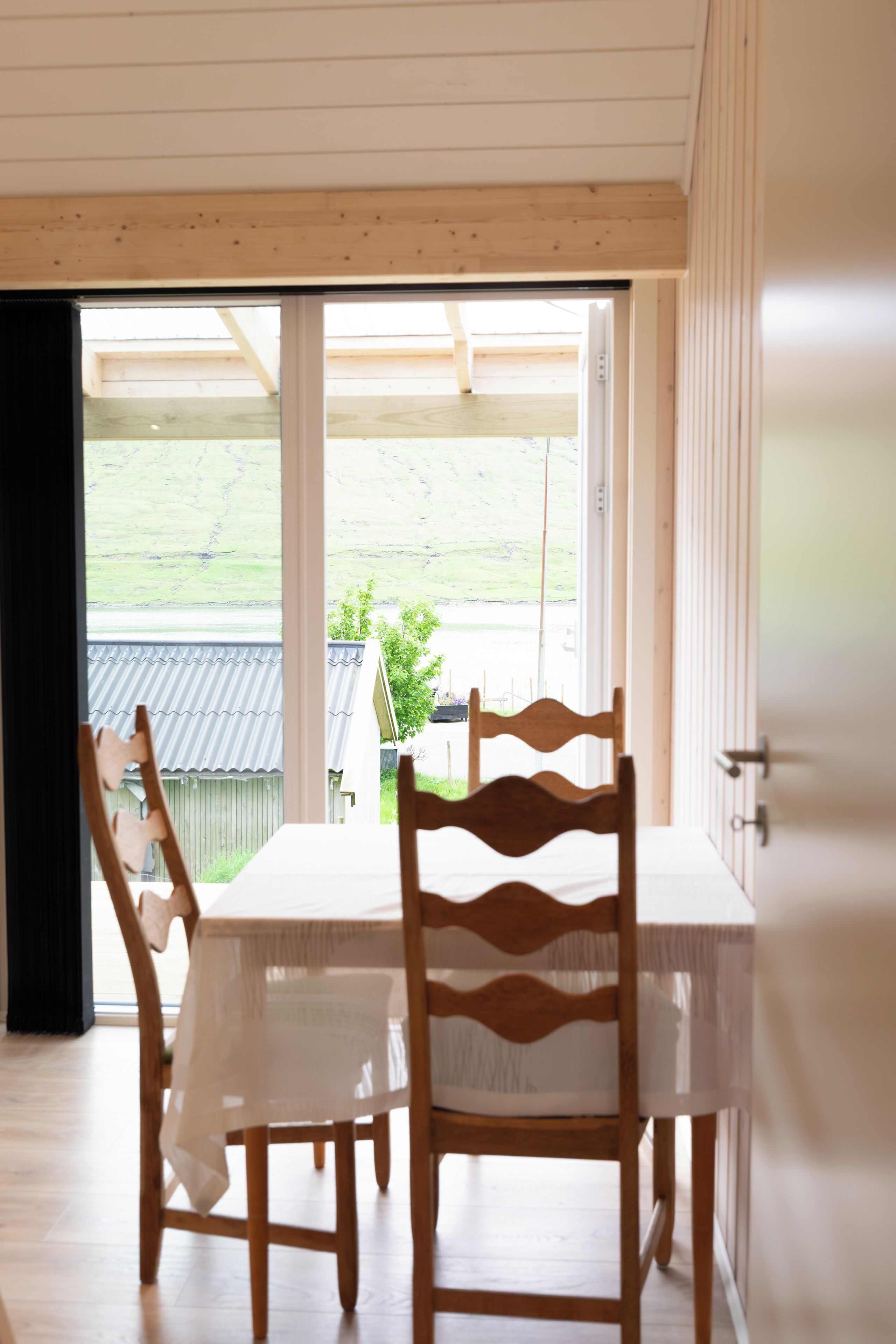 A dining area with a table and chairs, a view of a green landscape through a large window, and a wooden ceiling.