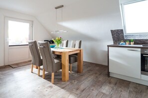 A modern kitchen with a dining area, featuring a wooden table, grey chairs, and a white cabinet with a built-in oven. - Holiday apartment with 2 bedrooms (Norderney)