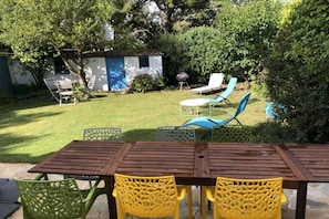 A patio with a wooden table and chairs, surrounded by greenery and a small shed. - Villa Les Brisants South Brittany (Loctudy)