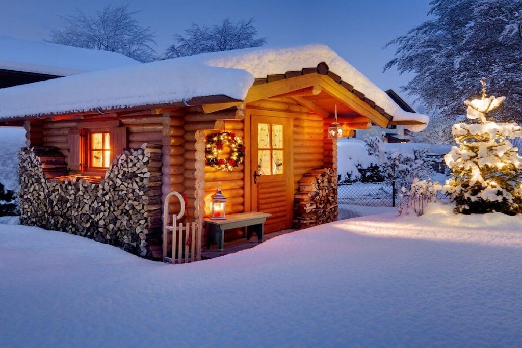 A log cabin with a snow-covered roof, a lit lantern, and a wreath on the door.