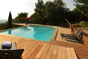 Eine Holzterrasse mit Schwimmbecken, Liegestühlen und einem roten Sonnenschirm.