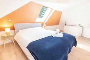 Et moderne soveværelse med skråvæg, ovenlys, en seng med blåt tæppe, et natbord med lampe og en hvid kommode.