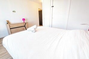 Een hotelkamer met een groot bed, een houten dressoir, een klein tafeltje met een lamp en een deur naar een andere kamer.
