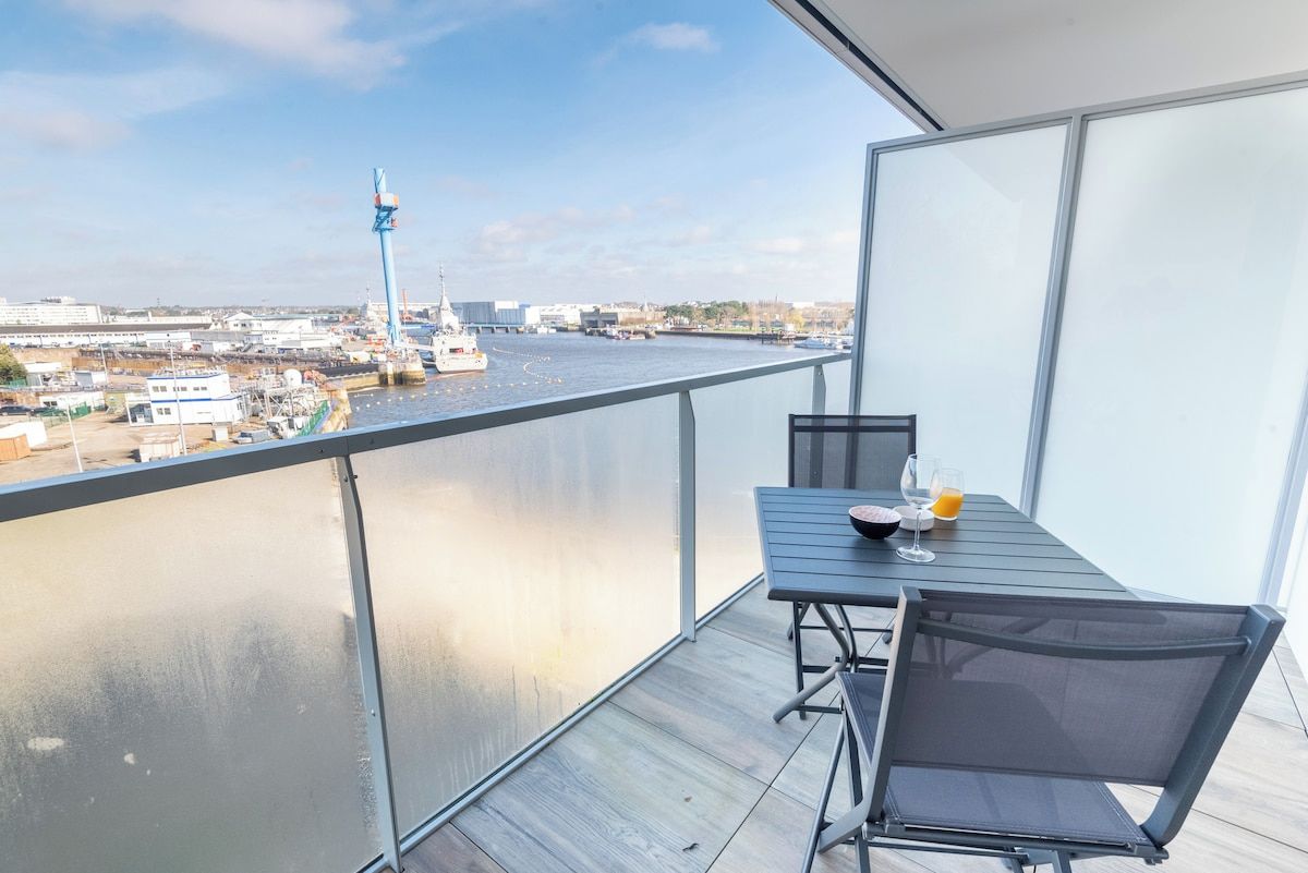 A balcony with a table and chairs, overlooking a body of water with a ship and industrial structures in the background.