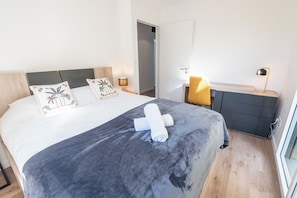 Ein ordentlich bezogenes Bett mit grauer Decke und aufgerollten Handtüchern, eine Nachttischlampe und ein kleiner Nachttisch mit einer Lampe.