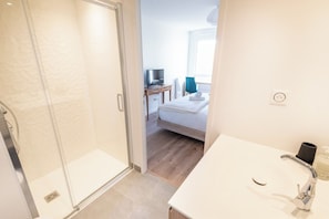 A hotel room with a shower, a bed, a bedside table with a TV, and a bathroom with a sink and mirror. - Premium 3-bedroom apartment with a view of Lorient Roadstead – by Groom* (Lorient)