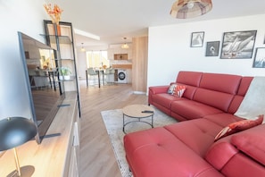 A modern living room with a red sofa, a coffee table, and a dining area with chairs and a table. - Premium 3-bedroom apartment with a view of Lorient Roadstead – by Groom* (Lorient)