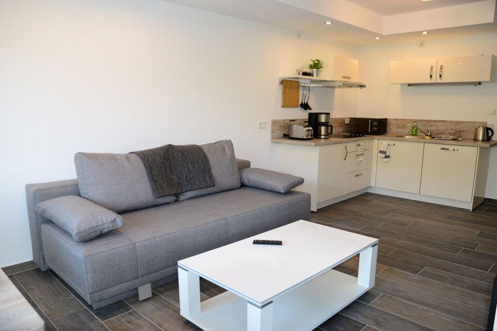Una sala de estar moderna con un sofá gris, una mesa de centro blanca y una cocina equipada con armarios y electrodomésticos.